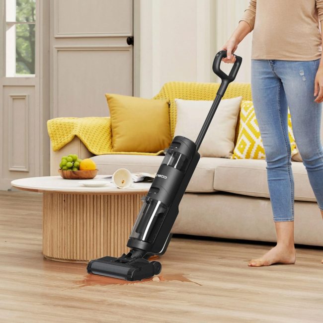 Person using a vacuum cleaner on a wooden floor with a spill near a coffee table and beige sofa