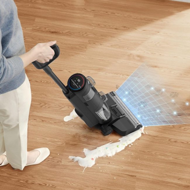 Person using a sensor-equipped cordless vacuum to clean a liquid spill on hardwood floor, featuring digital display