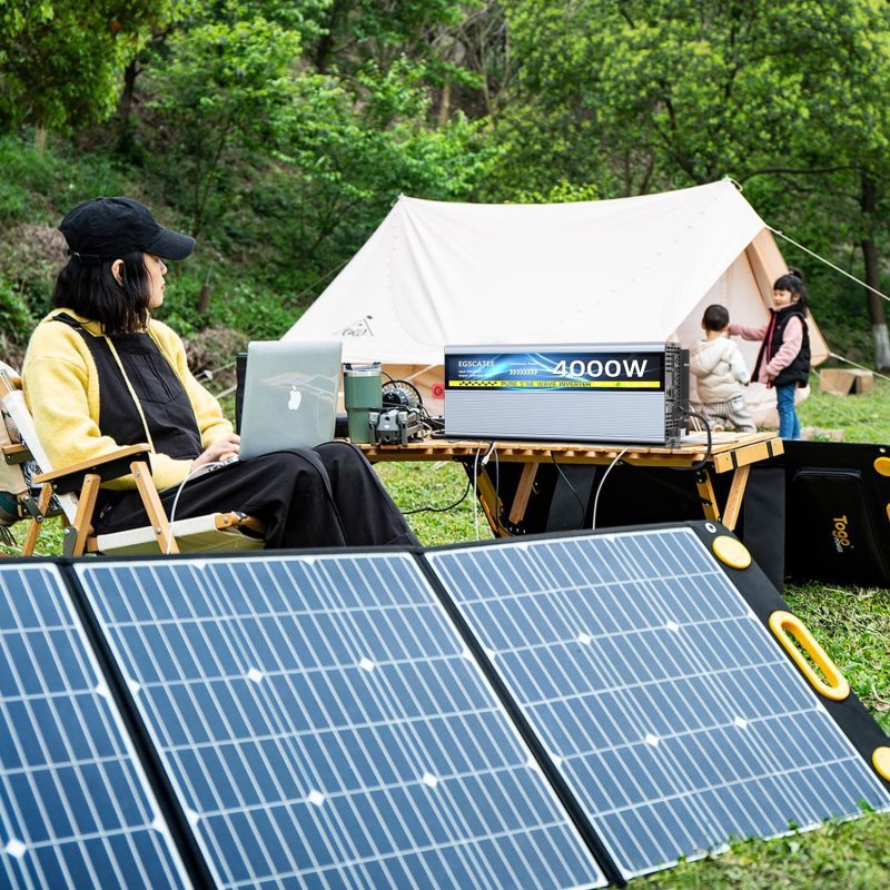 Person using a laptop at a campsite powered by solar panels and a 4000W inverter, with children near a tent in a wooded area.