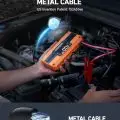 Orange battery jump starter with digital display showing 12.0V in car engine compartment