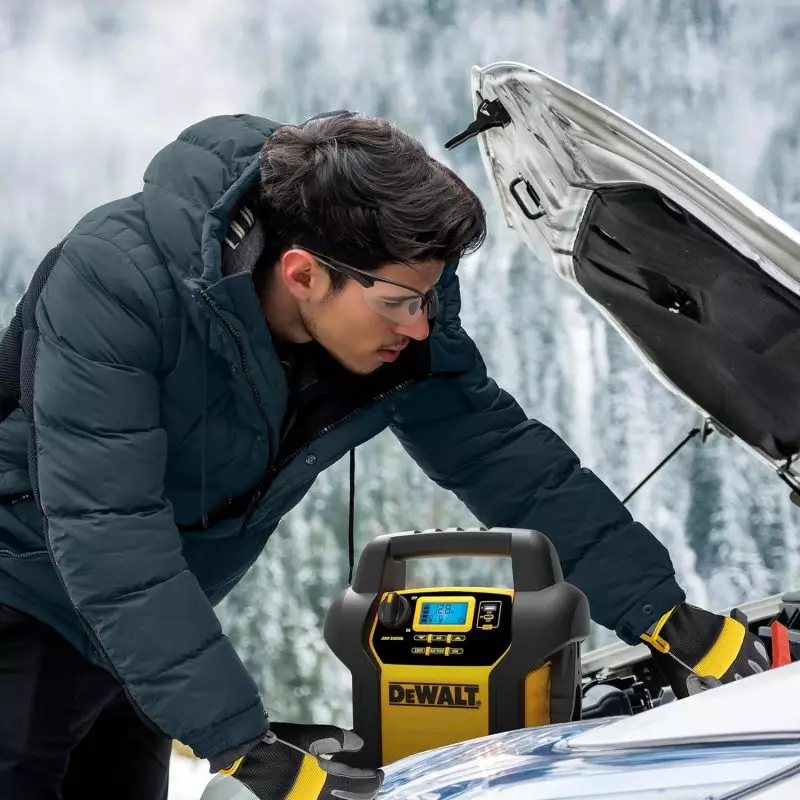 Mechanic in winter jacket and safety goggles operating a DEWAL Jump Starter on car battery in snowy environment