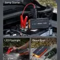 AstroAI multifunctional device being used as a jump starter connected to a car battery,   with built-in LED flashlight and charging smartphone
