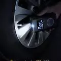 Person using a portable device to inflate car tire with digital display showing air pressure and LED light for visibility
